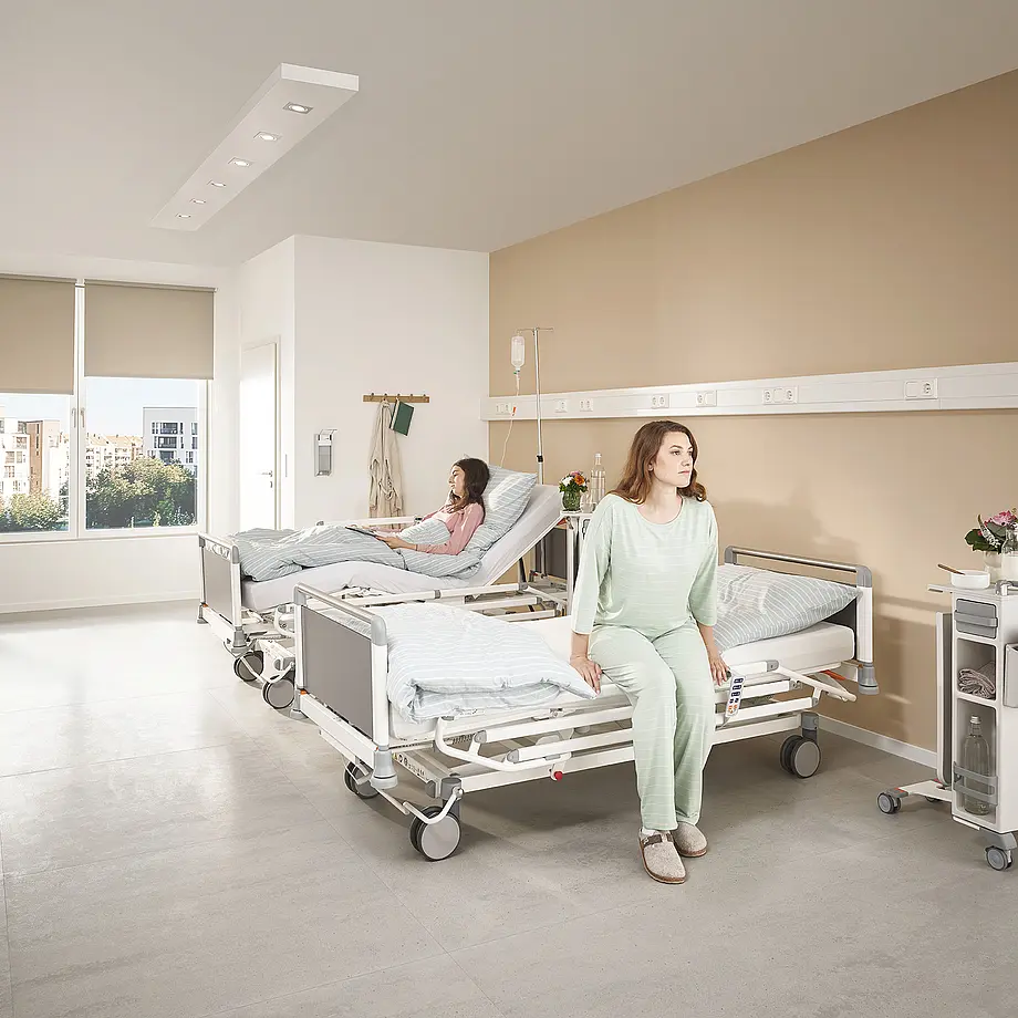 Patientin sitzt auf Evario one Pflegebett mit ergonomischer Aufstehhöhe im Klinikzimmer.